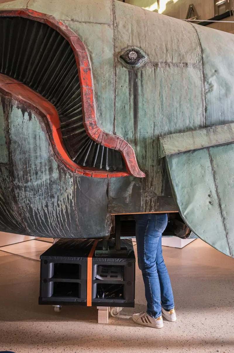 A visitor in jeans stands beside the 10-meter copper whale sculpture "Poldi," designed by artist Maria Benke, during the Wien Museum's Tage der Restaurierung (Days of Restoration) program in 2024. The weathered patina and red-painted lips of the massive artwork are visible, along with structural details and an orange-trimmed storage unit positioned beneath the sculpture. This piece was salvaged from the demolished Gasthaus zum Walfisch and represents a significant restoration project documenting the conservation process.