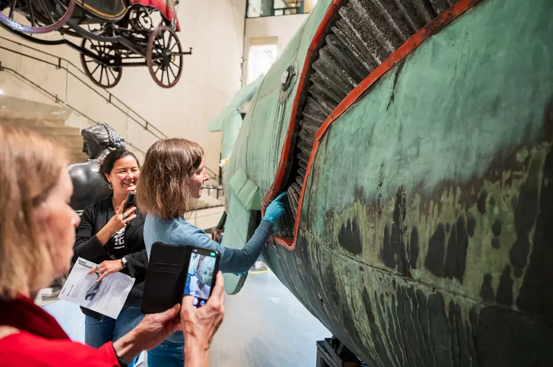 Museum visitors and staff examine the copper sheeting and patina of the 10-meter Prater whale sculpture 'Poldi' during the 'Tage der Restaurierung' program at the Wien Museum in 2024. A woman points to specific restoration features while attendees document the conservation work with smartphones, providing intimate access to details normally visible only to industrial climbers who maintain Maria Benke's design annually. The lowered sculpture allows guests to observe the craftsmanship and restoration techniques typically inaccessible to the public.