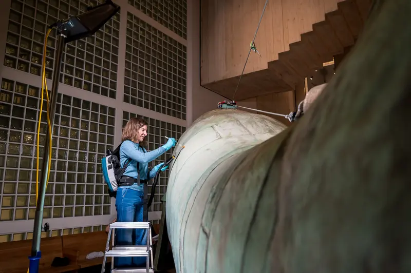 A conservator in blue work clothing examines the massive copper surface of Poldi, a 10-meter whale sculpture by Maria Benke, during the Wien Museum's Tage der Restaurierung (Conservation Days) program in 2024. The worker stands on a ladder to access the whale's curved body, demonstrating the monumental scale of this historic artwork salvaged from the demolished Gasthaus zum Walfisch in Vienna's Prater district. The industrial museum setting with metal grid walls and suspended wooden beams provides the backdrop for this behind-the-scenes conservation documentation.