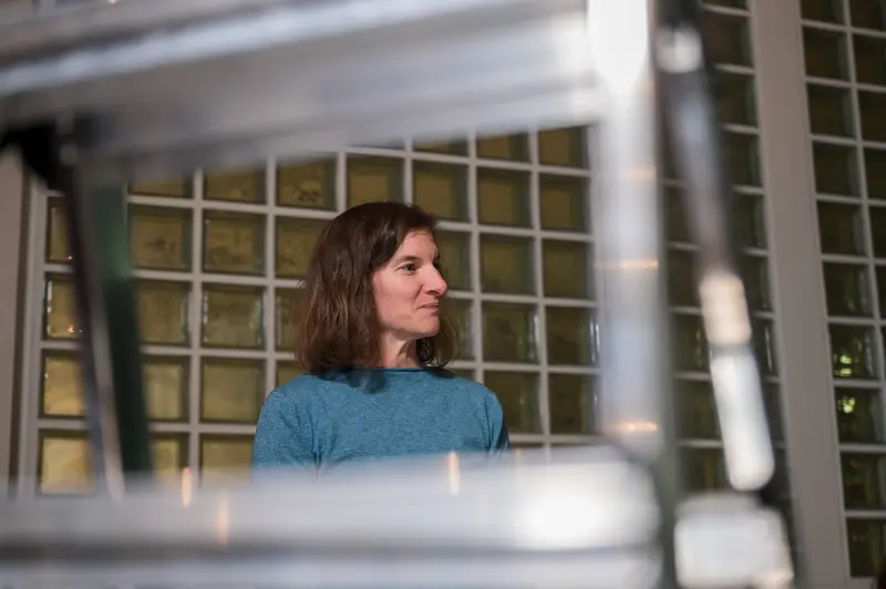 A museum conservator with shoulder-length brown hair wearing a teal sweater participates in the Wien Museum's 2024 Restoration Days program in Vienna, Austria. The professional is photographed through metal scaffolding framework, looking thoughtfully to the side while engaged in behind-the-scenes conservation work. The "Tage der Restaurierung" (Restoration Days) event showcases the specialized professionals responsible for preserving the Wien Museum's cultural artifacts and collections.