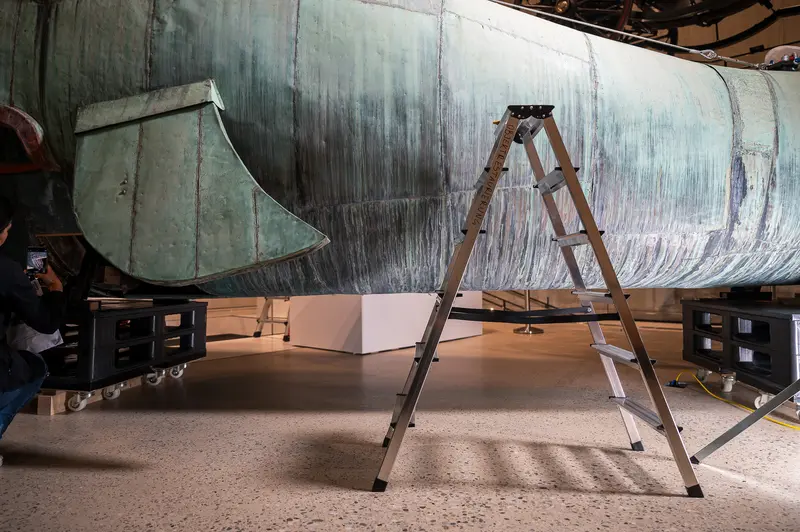 Museum conservators positioned a wooden step ladder beneath the copper-sheeted whale sculpture "Poldi" during the Wien Museum's restoration days, providing access for close examination of Maria Benke's rescued Prater artwork. The industrial conservation setting reveals behind-the-scenes restoration work during the "Tage der Restaurierung" program, with museum storage equipment visible in the background. The 10-meter sculpture underwent detailed conservation assessment in this Vienna facility.