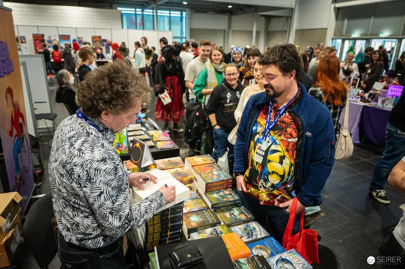 Book Signing at Vienna ComicCon 2024: Author Engaging with Fan at Comic Book Table