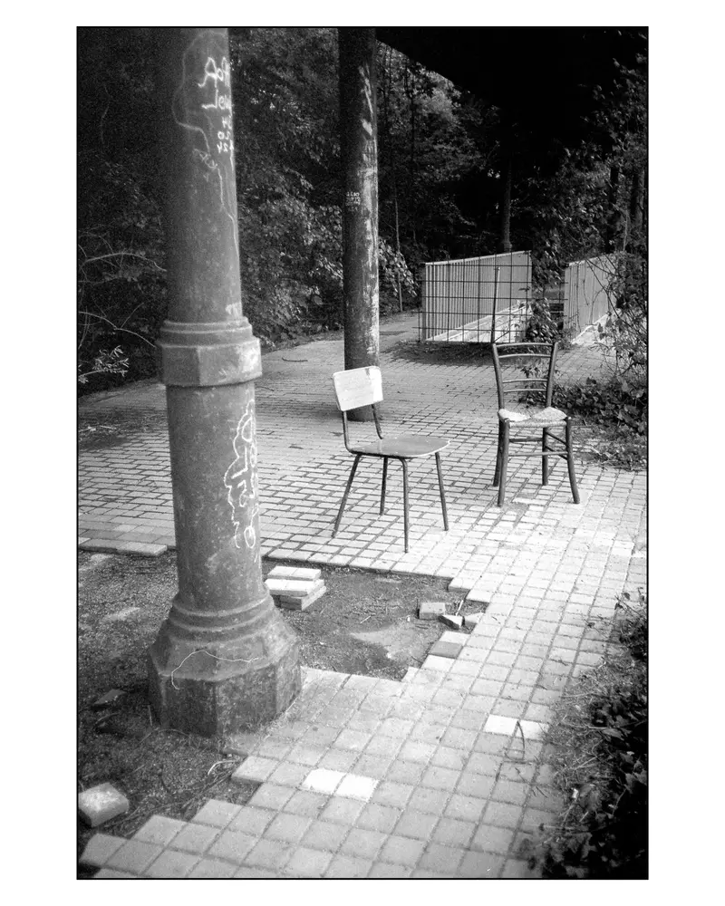 Two Abandoned Chairs on Cobblestone Path Beneath Graffitied Iron Pillar, Villach 2024