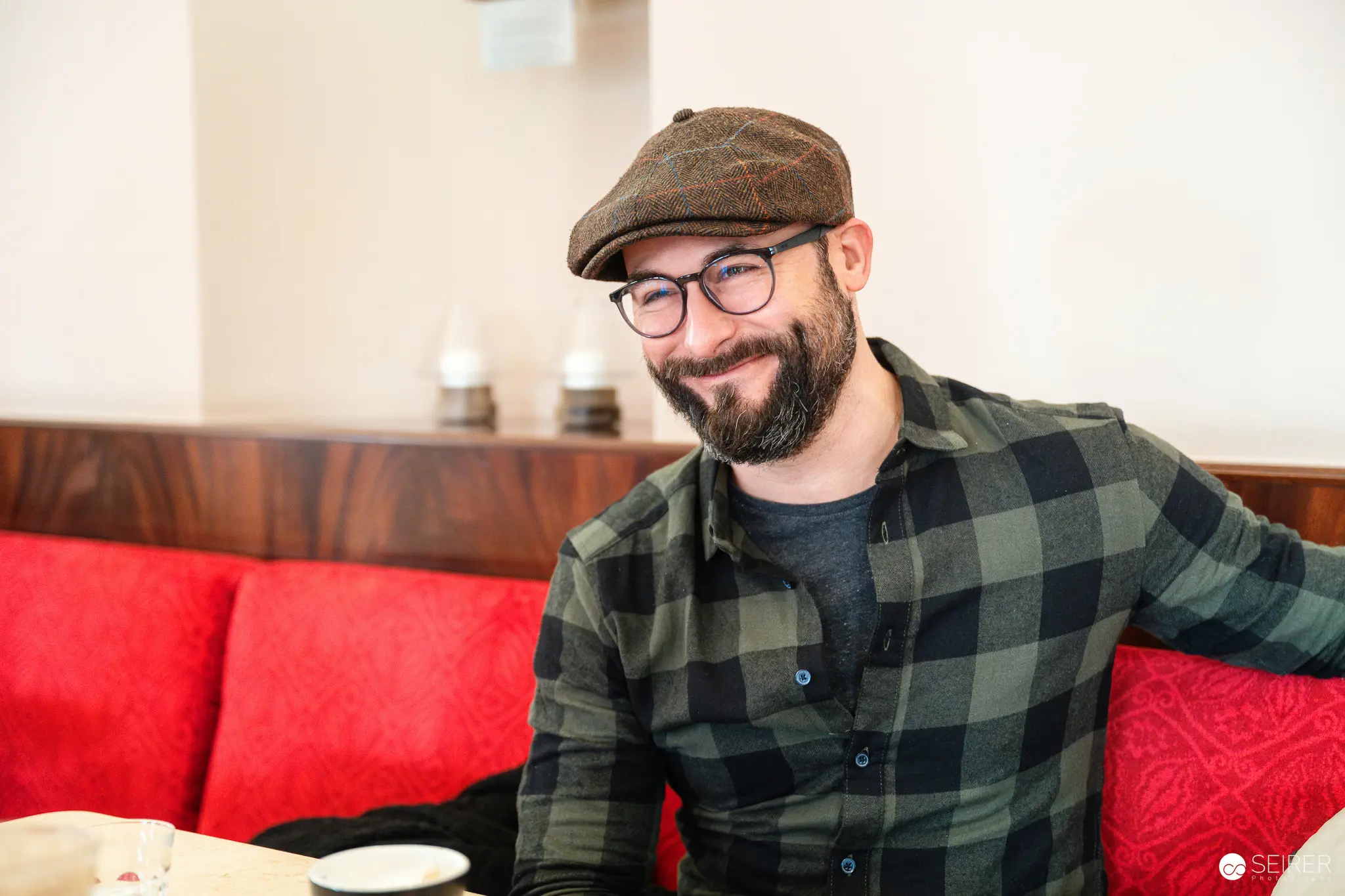 Smiling Bearded Man in Plaid Shirt and Flat Cap at Cafe