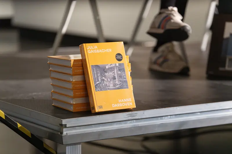 A stack of orange-spined copies of Julia Gaisbacher's photobook *Hanne Darboven. Am Burgberg* rests on a metallic surface in the foreground, with one volume positioned upright to display the cover featuring the artist's name and a black-and-white interior photograph. The shallow depth of field renders the background elements—including chair legs and footwear—softly out of focus, directing attention to the publication that serves as the central subject of this book presentation at FOTO ARSENAL WIEN. The controlled lighting and neutral tones emphasize the book's physical presence as both artistic object and documentary record of Gaisbacher's photographic investigation into Hanne Darboven's domestic and working spaces.