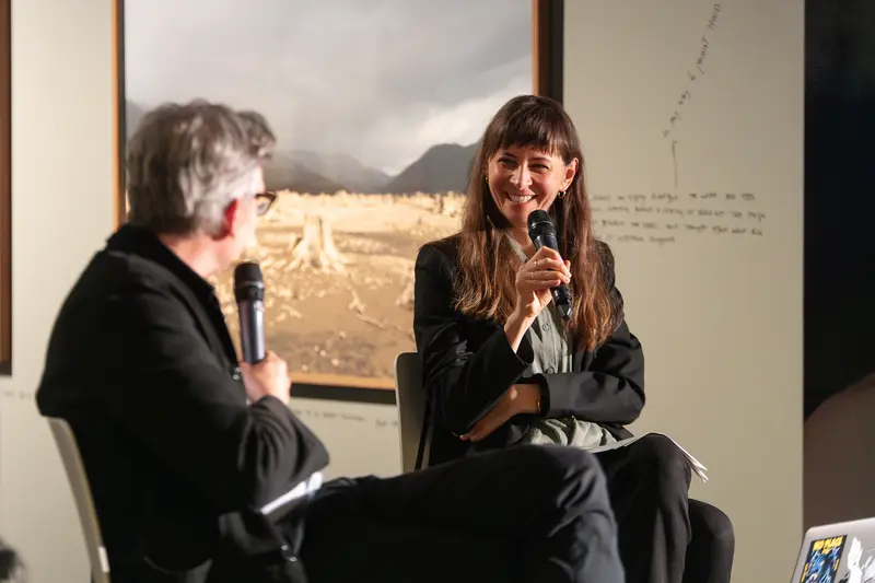 Felix Hoffmann and Julia Gaisbacher engage in animated dialogue during their book presentation at FOTO ARSENAL WIEN, both speakers holding microphones as they sit in profile against the gallery's white walls. The conversational dynamic is captured through their attentive postures and direct interaction, with Gaisbacher's expressive gesture suggesting active discussion of her photographic documentation of Hanne Darboven's Hamburg-Harburg spaces. The institutional setting's neutral backdrop and controlled lighting emphasize the scholarly nature of the discourse surrounding Gaisbacher's photobook *Hanne Darboven. Am Burgberg*.