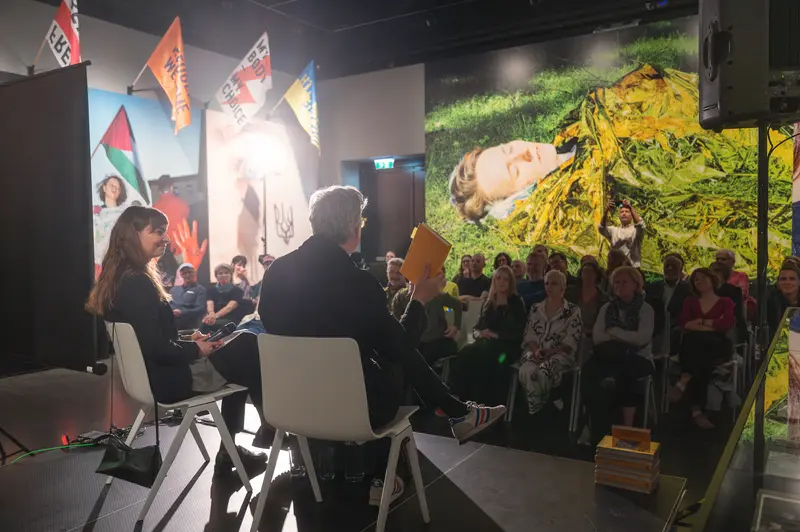 The book presentation continues at FOTO ARSENAL WIEN with Felix Hoffmann, Artistic Director, and Julia Gaisbacher seated in white contemporary chairs before an audience, engaged in dialogue about Gaisbacher's photobook *Hanne Darboven. Am Burgberg*. The gallery's controlled lighting illuminates the conversational setting while maintaining focus on the speakers, with Hoffmann gesturing as he addresses questions or commentary about the multi-year photographic documentation of Darboven's Hamburg-Harburg spaces. The composition captures the intimate scale of the institutional talk format, positioning the discussion within the broader context of contemporary photographic discourse and archival practice.