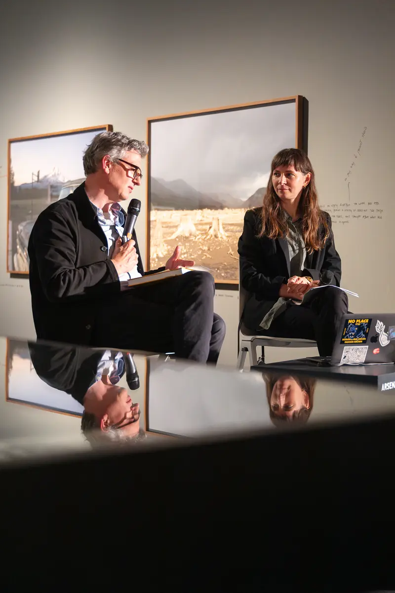 Felix Hoffmann, Artistic Director of FOTO ARSENAL WIEN, and photographer Julia Gaisbacher engage in dialogue during the presentation of her photobook *Hanne Darboven. Am Burgberg*, seated on modern chairs within the gallery's minimalist white-walled space. The compositional arrangement captures both speakers in profile against the neutral institutional backdrop, with Hoffmann gesturing toward Gaisbacher as she holds materials related to her documentation project, their positioning suggesting active scholarly exchange about her photographic investigation of the conceptual artist's Hamburg-Harburg residences. The controlled lighting and reflective flooring characteristic of the contemporary gallery environment frame this moment of cultural discourse within FOTO ARSENAL WIEN's exhibition space.