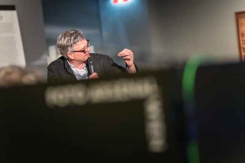 A speaker with glasses and graying hair, wearing a dark jacket over a light shirt, gestures with raised hand while holding a microphone during the book presentation at FOTO ARSENAL WIEN. The figure is captured mid-speech against the gallery's neutral walls, with dramatic overhead lighting creating strong contrast and selective focus that isolates the speaker from the softly blurred background. The composition emphasizes the dynamic nature of the discourse surrounding Gaisbacher's photographic documentation of Hanne Darboven's Hamburg-Harburg spaces.