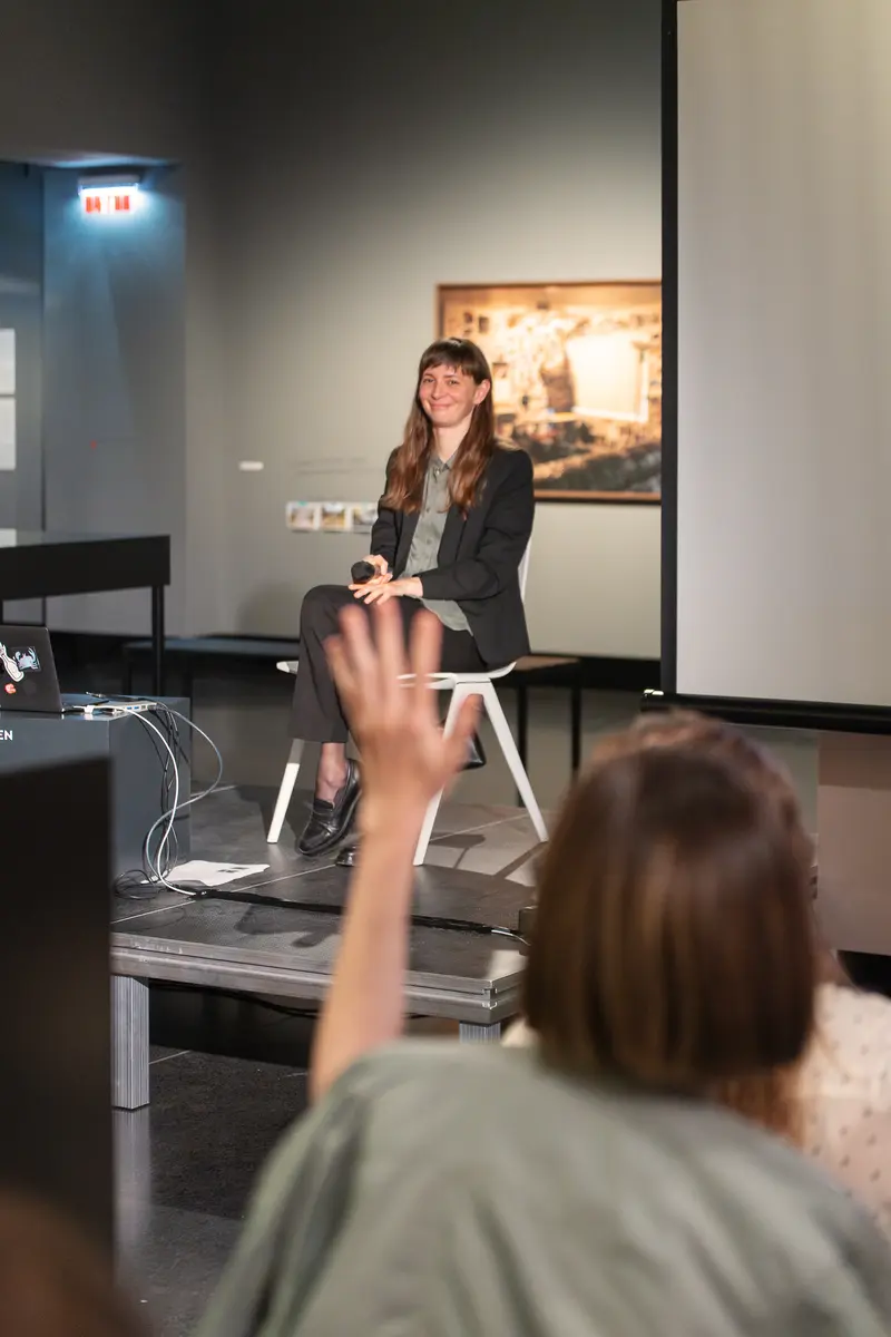 A member of the audience raises their hand during Julia Gaisbacher's presentation of her photobook *Hanne Darboven. Am Burgberg* at FOTO ARSENAL WIEN, with Gaisbacher visible in the background seated against the gallery's white wall. The composition captures the interactive nature of the book talk, showing the dialogue between presenter and attendees in the institutional setting of the photography center. The controlled gallery lighting and neutral backdrop emphasize the scholarly discourse surrounding Gaisbacher's photographic documentation of the conceptual artist's Hamburg-Harburg residences.