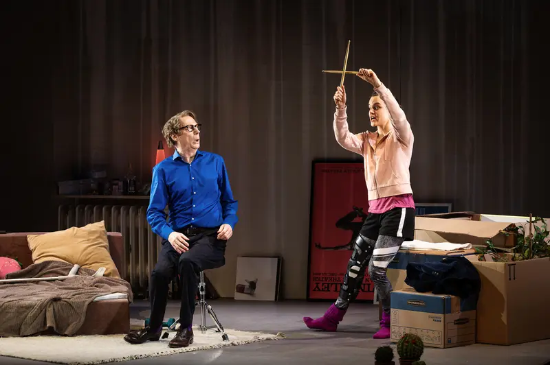 A scene from the dress rehearsal of "Die Tanzstunde" at Kammerspiele Wien shows André Pohl as Professor Ever Montgomery observing Katharina Klar as dancer Senga Quinn, who stands elevated with arms raised holding drumsticks in a conducting gesture. The staging, designed by Stephan von Wedel, features a domestic interior with a bed strewn with cushions at left, cardboard moving boxes, and a piano partly visible at right, all set against heavy draped curtains under Sebastian Schubert's lighting design. The composition emphasizes the vertical relationship between the two performers, with Montgomery positioned at ground level in bright blue shirt while Quinn commands the space from above in layered pink and purple athletic wear.