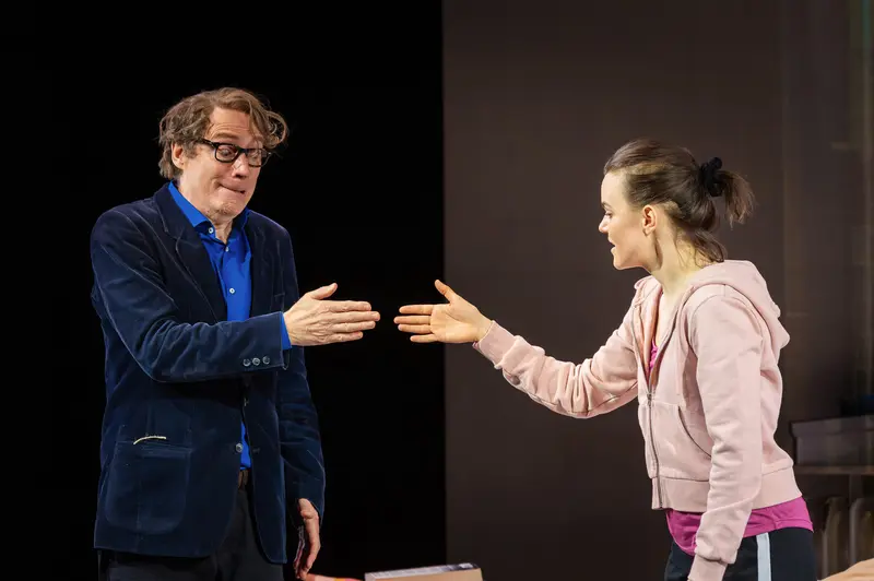 In a photographed dress rehearsal scene from "Die Tanzstunde" at Kammerspiele Wien, actors André Pohl and Katharina Klar perform a moment of tentative physical connection against a stark divided backdrop of black and warm brown tones. Pohl, positioned stage left in a navy velvet blazer and blue shirt, extends his hands palm-down toward Klar, who mirrors the gesture from stage right in a pale pink hoodie and magenta athletic wear, her hair secured in a tight bun. The symmetrical hand positioning and the actors' focused eye contact suggest a choreographed teaching moment between Pohl's character, Professor Ever Montgomery, and Klar's dancer Senga Quinn, lit with precise theatrical illumination designed by Sebastian Schubert.