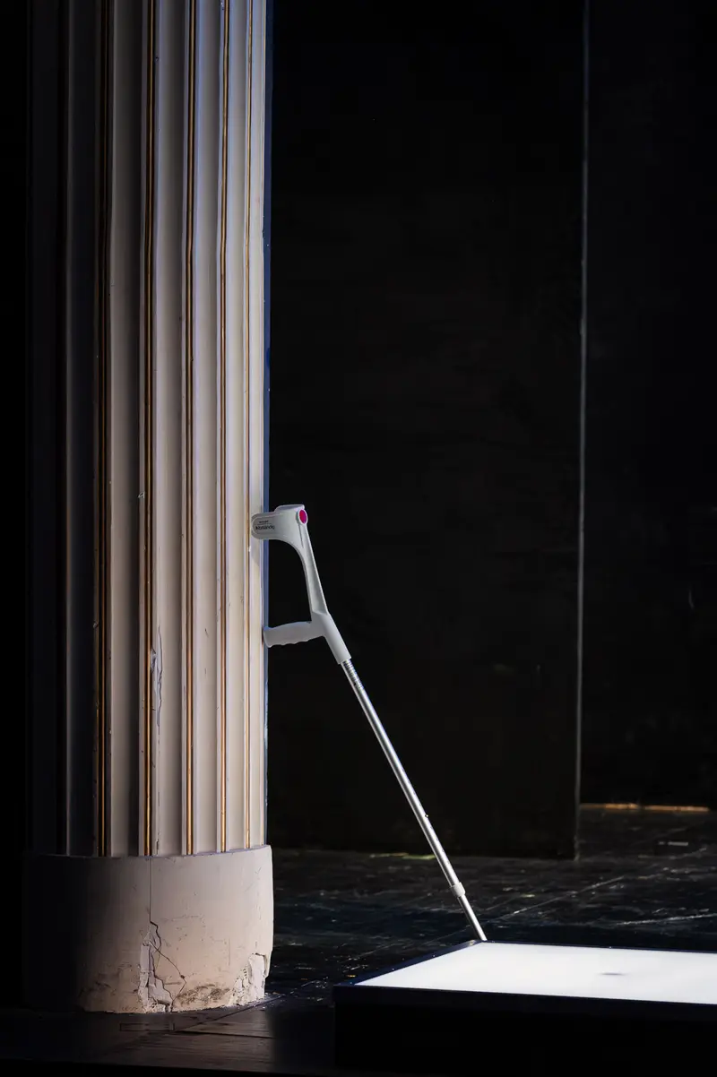 A single crutch stands illuminated against the stark black floor and void of the stage, positioned adjacent to a classical fluted column bathed in cool white light. The composition captures the minimalist scenic design by Stephan von Wedel for "Die Tanzstunde" at Kammerspiele Wien, where the austere architectural element and medical aid create a formal tension between permanence and fragility. Directed by Folke Braband, this production features Katharina Klar as dancer Senga Quinn and André Pohl as Professor Ever Montgomery, with the isolated prop suggesting themes of physical limitation and theatrical absence within the performance space photographed during technical rehearsal on January 14, 2026.