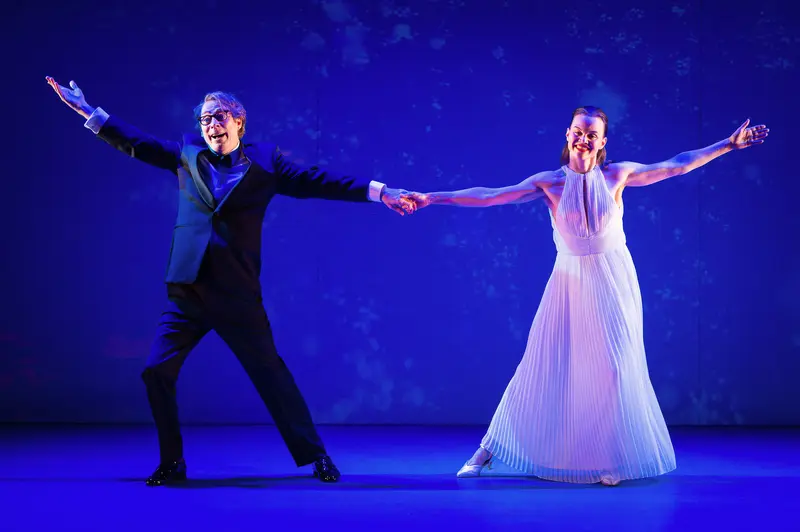 Katharina Klar and André Pohl perform a ballroom dance sequence against a saturated blue backdrop during a photographic rehearsal of "Die Tanzstunde" at Kammerspiele Wien. The actors extend their outer arms in symmetrical gestures while maintaining hand contact at center, Pohl in dark formal attire and Klar in a flowing, pleated white gown that catches the stage lighting. The composition emphasizes theatrical geometry through the performers' parallel diagonal stances and the stark tonal contrast between costumes, while atmospheric lighting creates a nocturnal ambiance characteristic of Stephan von Wedel's scenic design and Sebastian Schubert's illumination work for this production under Folke Braband's direction.