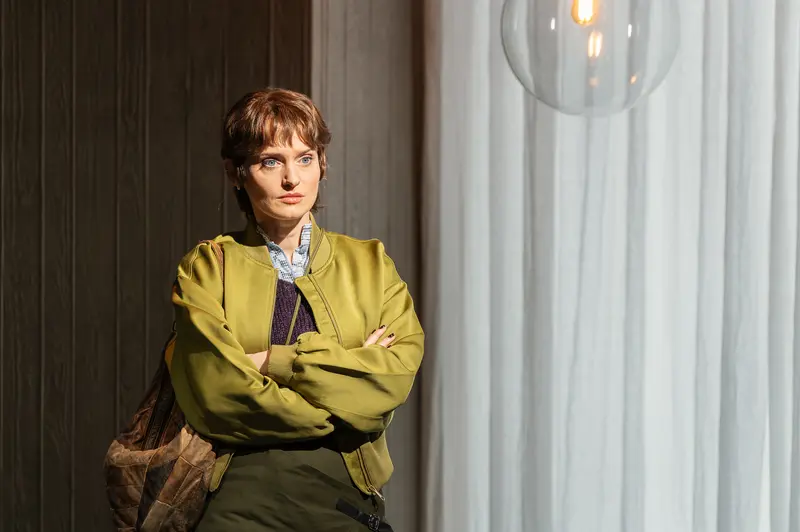 An actor stands with crossed arms wearing a yellow jacket over a collared shirt during a performance of Sophia at the Kammerspiele der Josefstadt in Vienna. The setting features white curtains and dark wood paneling with overhead pendant lighting. The photograph was taken by Michael Seirer Photography during the February 23, 2026 performance.