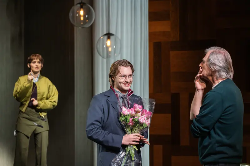 An actor wearing glasses and holding a bouquet of pink flowers stands center stage during a curtain call at the Kammerspiele der Josefstadt in Vienna. Two other performers are visible on stage, one wearing yellow attire to the left and another figure to the right, with hanging globe light fixtures and draped curtains forming the backdrop. The Kammerspiele der Josefstadt is a historic theater in Vienna's eighth district, known for producing classical and contemporary drama productions.