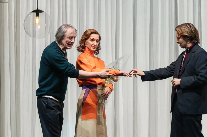 Three performers enact a scene from the production Sophia at the Kammerspiele der Josefstadt in Vienna. An older man in a teal sweater and a woman in an orange blouse hand documents to a younger man in a dark suit against white curtains forming the stage backdrop. The Kammerspiele der Josefstadt is a historic theater in Vienna's eighth district, Austria.