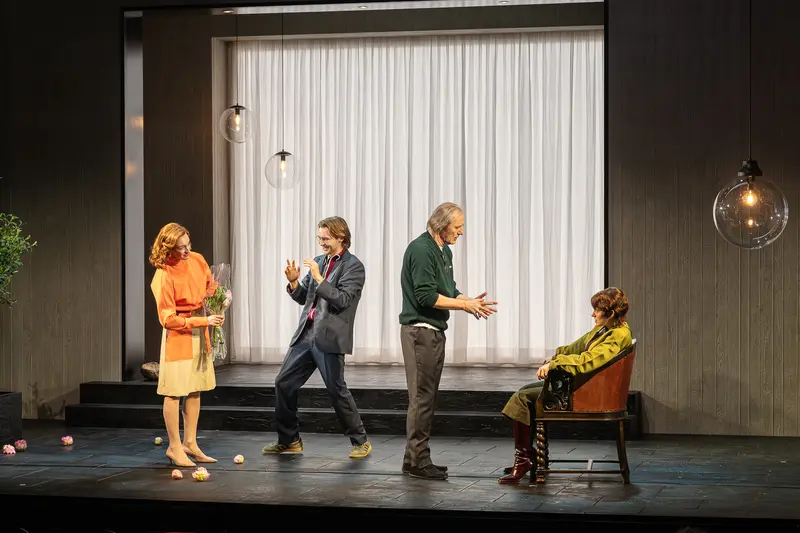 Four performers occupy a minimalist stage at the Kammerspiele der Josefstadt in Vienna, with a white curtained backdrop and dark walls. Three standing actors in an orange dress, blue suit, and green cardigan interact with a fourth performer seated in a brown leather chair, while scattered spherical objects lie on the stage floor. The Kammerspiele der Josefstadt is a historic chamber theater in Vienna's eighth district, known for intimate dramatic productions.