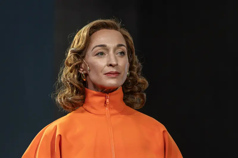 An actor performs in the production Sophia at the Kammerspiele der Josefstadt in Vienna during a February 23, 2026 performance. The performer wears a bright orange zip-up jacket and is photographed against a dark background with shoulder-length curled hair. The Kammerspiele der Josefstadt is a historic theater in Vienna's eighth district known for intimate dramatic productions.