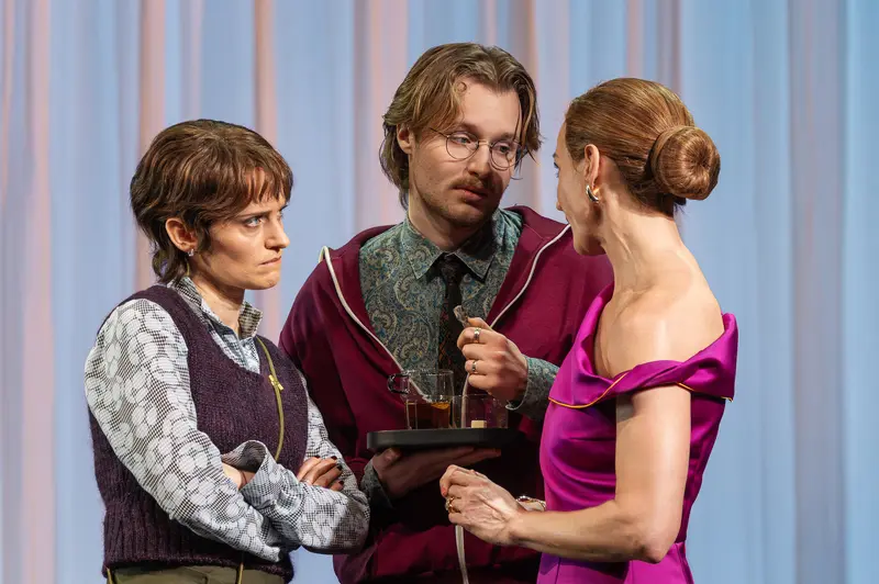 Three performers in period attire engage in a scene during a production of Sophia at the Kammerspiele der Josefstadt in Vienna. The left actor wears a patterned vest over a printed shirt, the center actor holds a tablet while wearing glasses and a burgundy robe, and the right actor appears in a magenta off-shoulder dress. The Kammerspiele der Josefstadt is a historic theater in Vienna's eighth district, known for intimate dramatic productions.