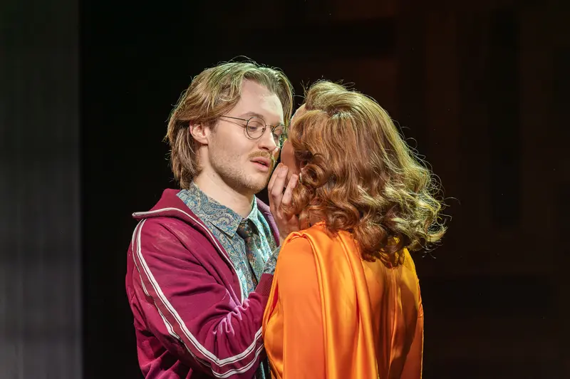 Two performers share a close theatrical moment during a production of Sophia at the Kammerspiele der Josefstadt in Vienna. The actor on the left wears glasses and a burgundy track jacket over a patterned shirt, while the performer on the right wears an orange garment, with both figures positioned in an intimate exchange on a darkened stage. The Kammerspiele der Josefstadt is a historic theater in Vienna's eighth district known for contemporary dramatic productions.