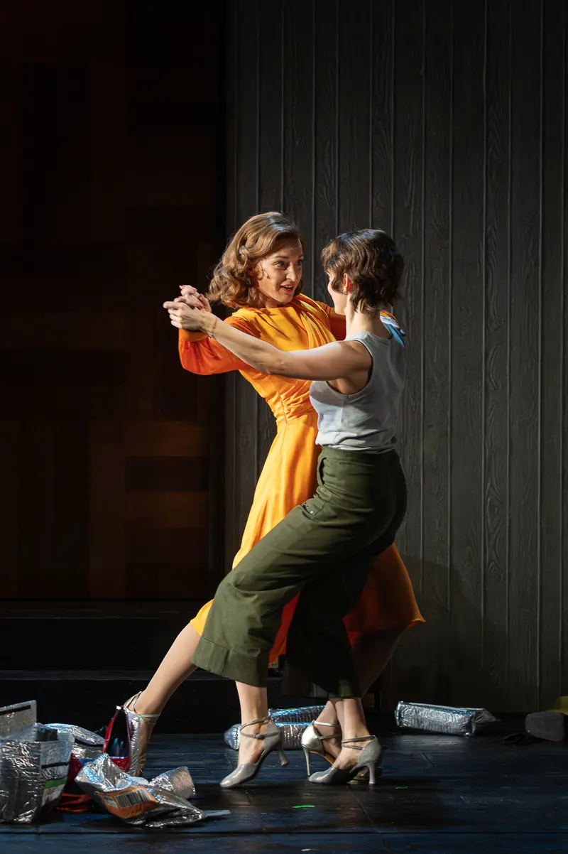 Two performers dance in intimate close embrace during a theatrical stage production, with one wearing a flowing yellow and orange dress and the other in olive green pants and a light-colored top. Both wear character shoes on a dark stage floor scattered with props, performing against dark paneled walls. The scene depicts a choreographed moment of connection typical of contemporary theatrical dance productions.