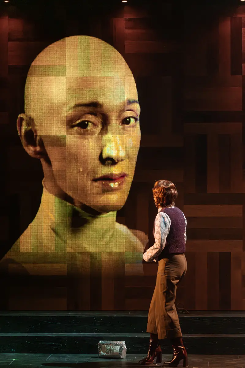 An actor performs on stage next to a large projected pixelated human face during a production of Sophia at the Kammerspiele der Josefstadt in Vienna. The performer wears casual clothing while positioned adjacent to the digitally rendered facial projection on a dark stage set. The production integrates live theatrical performance with digital imagery to create a hybrid visual experience.