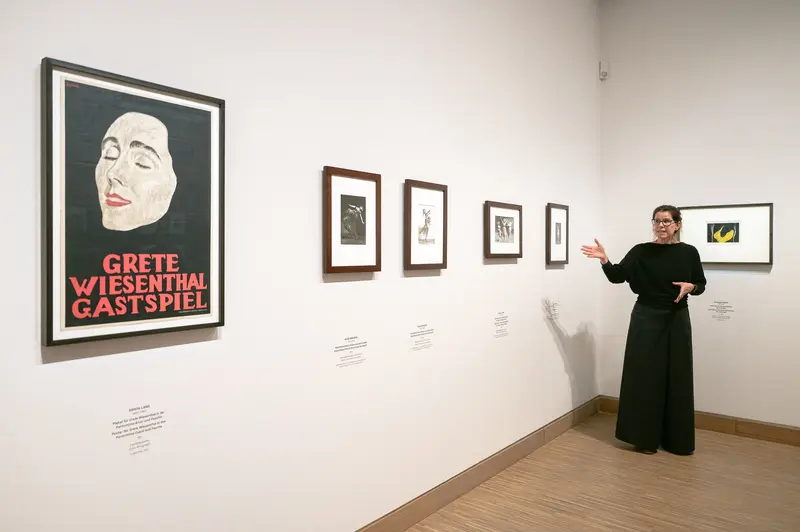 Astrid Mahler, curator of the Tanzbild exhibition at Albertina Modern in Vienna, points toward framed artworks displayed on white gallery walls in April 2026. A prominent "Grete Wiesenthal Gastspiel" poster featuring a stylized female profile in red and black dominates the left wall, accompanied by a series of smaller framed pieces arranged horizontally. The installation presents early 20th-century performance and theatrical promotional materials documenting Grete Wiesenthal's guest performances.
