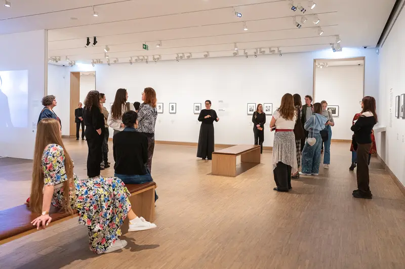 Visitors attend the Tanzbild exhibition at Albertina Modern in Vienna, April 2026, observing framed artworks displayed on white gallery walls while standing near wooden display pedestals in a minimalist, professionally lit space. Multiple attendees in casual and formal attire move throughout the gallery, engaging directly with individual contemporary artworks arranged across the expansive wooden floor. The spacious gallery environment is designed to facilitate visitor interaction with the featured contemporary art collection.