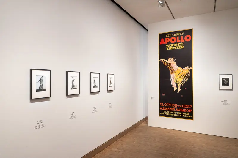 The Tanzbild exhibition at Albertina Modern in Vienna displays archival materials from the Apollo Theater, a historic New York City venue that opened in 1934 and became a cultural institution for African American entertainment. A vintage Apollo Theater variety playbill featuring a golden rooster illustration is mounted alongside four framed black-and-white performance photographs on a white gallery wall. The Apollo Theater's significant role in American performance history is documented through these exhibition materials.