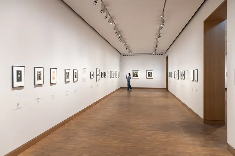 A visitor views framed drawings and prints displayed in the Tanzbild exhibition at Albertina Modern in Vienna, April 2026. Approximately thirty artworks are arranged in salon-style configurations across white walls under professional track lighting, with a wooden floor defining the minimalist gallery space. The installation represents a contemporary presentation format for curated exhibitions of works on paper in an institutional museum setting.