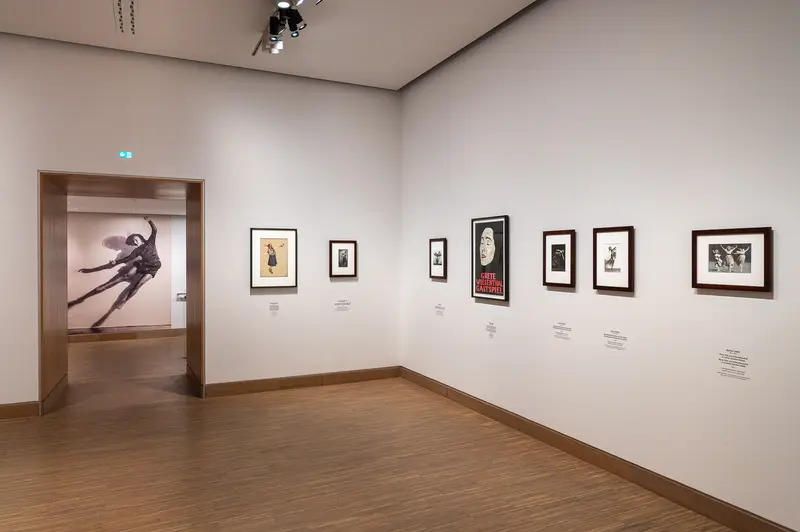 The Tanzbild exhibition at Albertina Modern in Vienna presents a curated selection of framed artworks and prints arranged salon-style across white gallery walls, with a sculptural installation visible through an open doorway. Multiple works of varying sizes, each accompanied by identifying plaques, are displayed on wooden flooring in a professional museum setting. The exhibition demonstrates the institutional presentation of visual art within Vienna's contemporary art institutions.