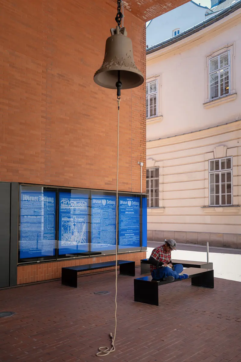 Person Seated Beneath Suspended Bell in Courtyard Installation