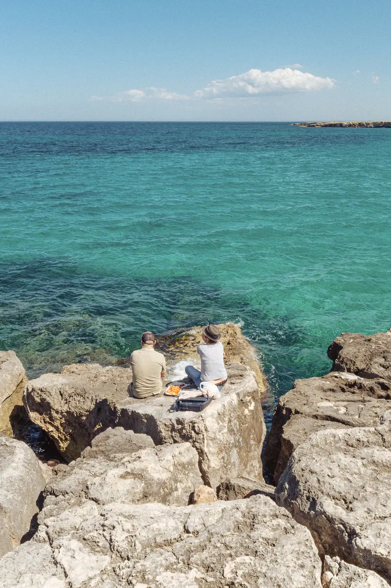 Two people sitting on rocky outcrop overlooking turquoise Mediterranean sea