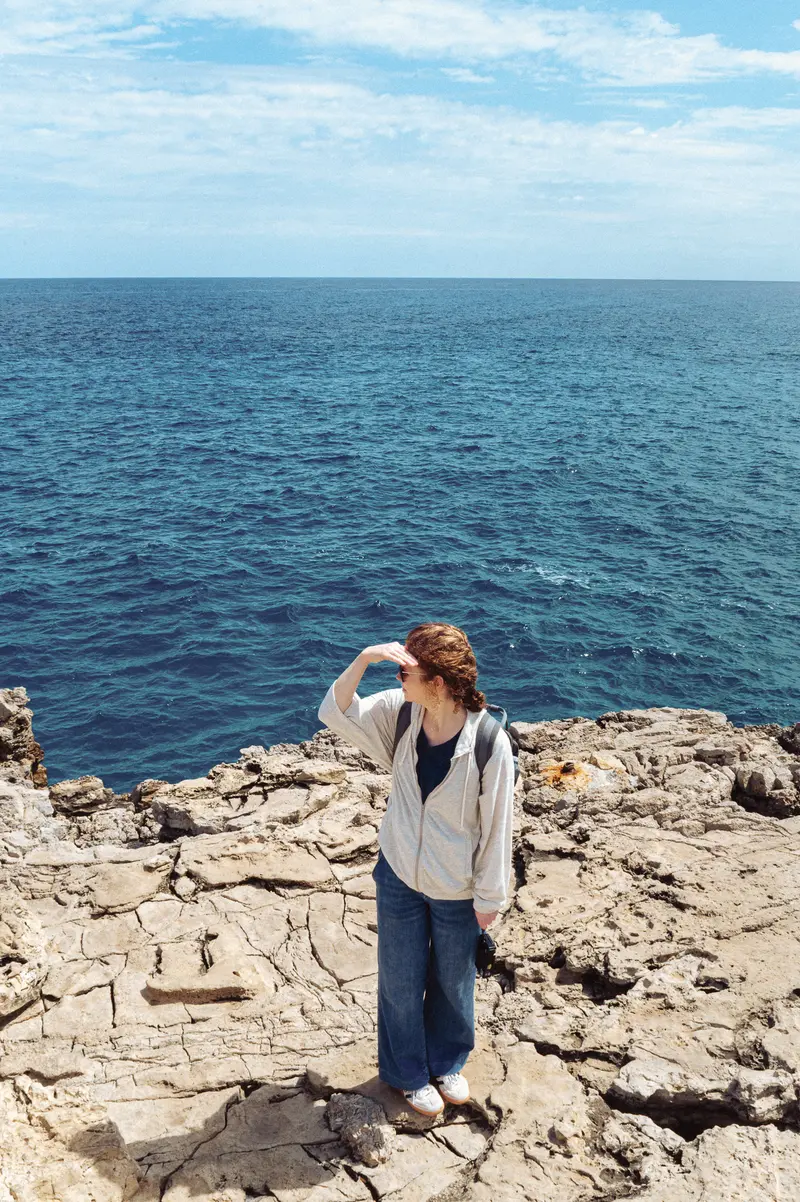 Woman in White Cardigan Standing on Rocky Coastal Cliff Overlooking Sea