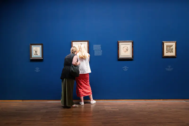 Two Visitors Examining Framed Drawings on Deep Blue Gallery Wall