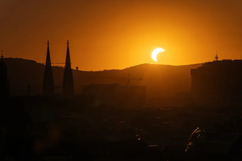 Partially eclipsed sun setting behind Vienna Woods above Votivkirche twin spires, 12 August 2026